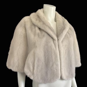 May include: A short-sleeved, cropped bolero jacket made of soft, off-white faux fur. The garment features a wide, folded shawl collar and a clean, open front design that sits at the waist, providing a textured, plush appearance throughout the piece.