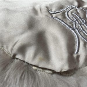 May include: Close-up of a cream-colored, possibly vintage, fur accessory. The lining is a light satin with embroidered silver-gray script. The fur appears soft and fluffy.