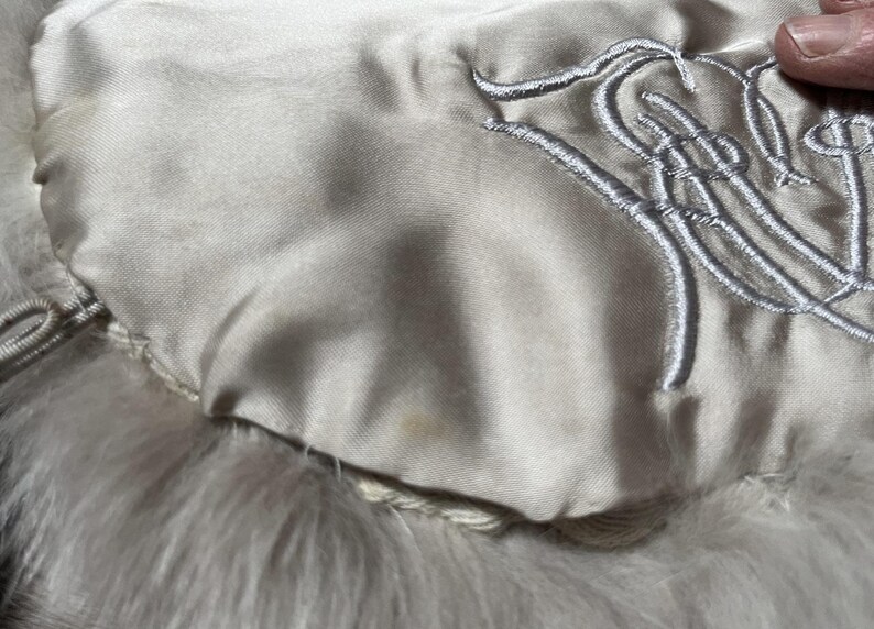 May include: Close-up of a cream-colored, possibly vintage, fur accessory. The lining is a light satin with embroidered silver-gray script. The fur appears soft and fluffy.