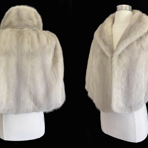 May include: A light grey fur capelet featuring a wide shawl collar and short sleeves. The garment is displayed on a mannequin, showing both the back view with its rounded hem and the front view with an open, draped neckline and soft texture.