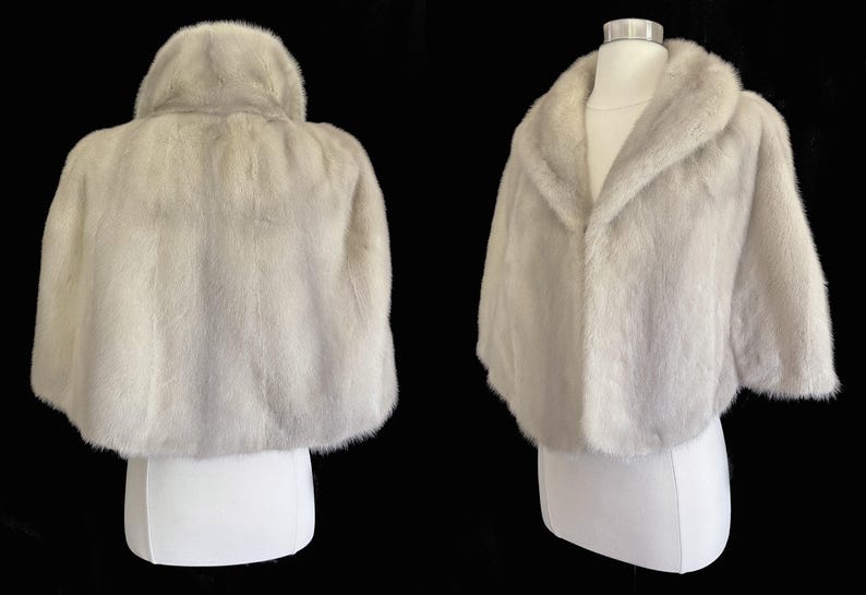 May include: A light grey fur capelet featuring a wide shawl collar and short sleeves. The garment is displayed on a mannequin, showing both the back view with its rounded hem and the front view with an open, draped neckline and soft texture.