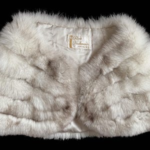 May include: A light gray fur bolero with a satin lining. The bolero has a rounded shape and a label that reads "Real Nachmans Fur Salon". The fur appears soft and luxurious, with a natural texture.
