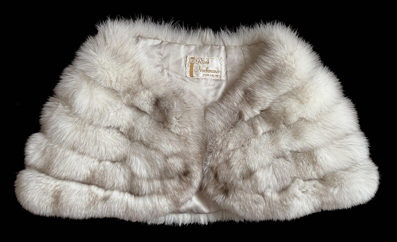 May include: A light gray fur bolero with a satin lining. The bolero has a rounded shape and a label that reads "Real Nachmans Fur Salon". The fur appears soft and luxurious, with a natural texture.