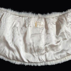 May include: A vintage white fur stole with a satin lining. The stole has a curved shape, with a fluffy fur trim around the edges. The interior is a smooth, off-white satin. A small embroidered design is visible on the left side.