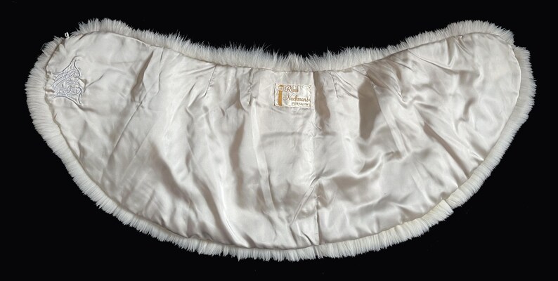 May include: A vintage white fur stole with a satin lining. The stole has a curved shape, with a fluffy fur trim around the edges. The interior is a smooth, off-white satin. A small embroidered design is visible on the left side.