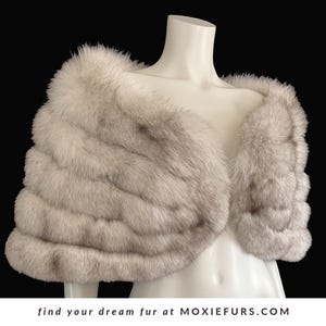 May include: A light gray fur stole displayed on a mannequin. The stole has a layered design and a soft, textured appearance. The text "find your dream fur at MOXIEFURS.COM" is at the bottom of the image.