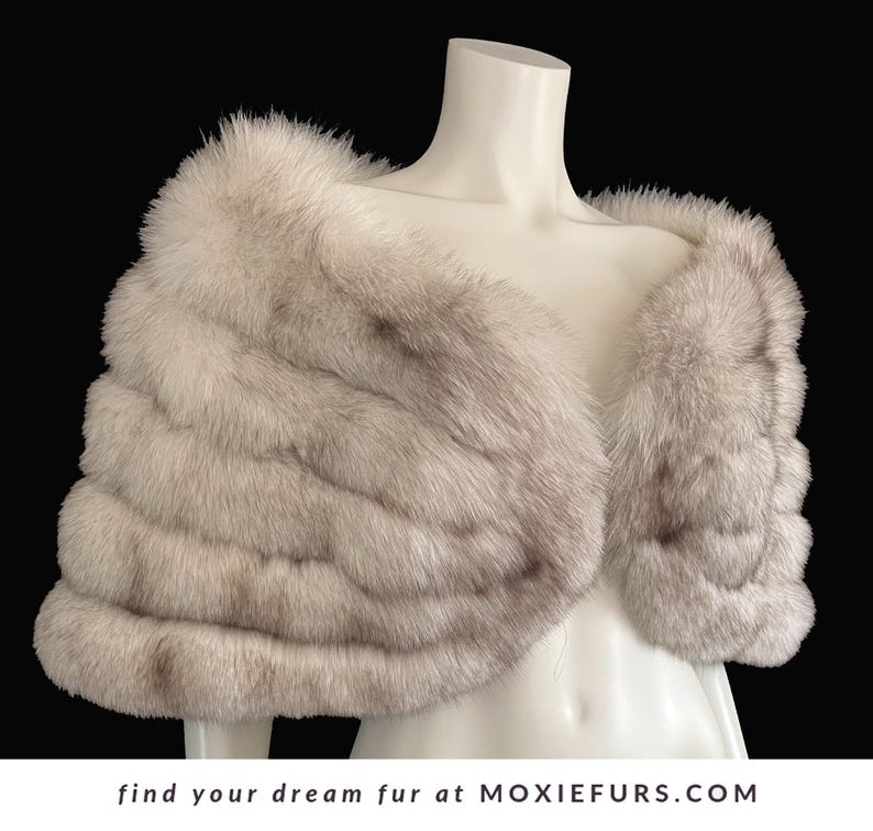 May include: A light gray fur stole displayed on a mannequin. The stole has a layered design and a soft, textured appearance. The text "find your dream fur at MOXIEFURS.COM" is at the bottom of the image.