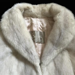 May include: A cream-coloured fur coat with a wide collar and a smooth, light-coloured satin lining. A rectangular fabric label is sewn into the interior neck area, featuring the text Steiger's and additional smaller, partially visible text.