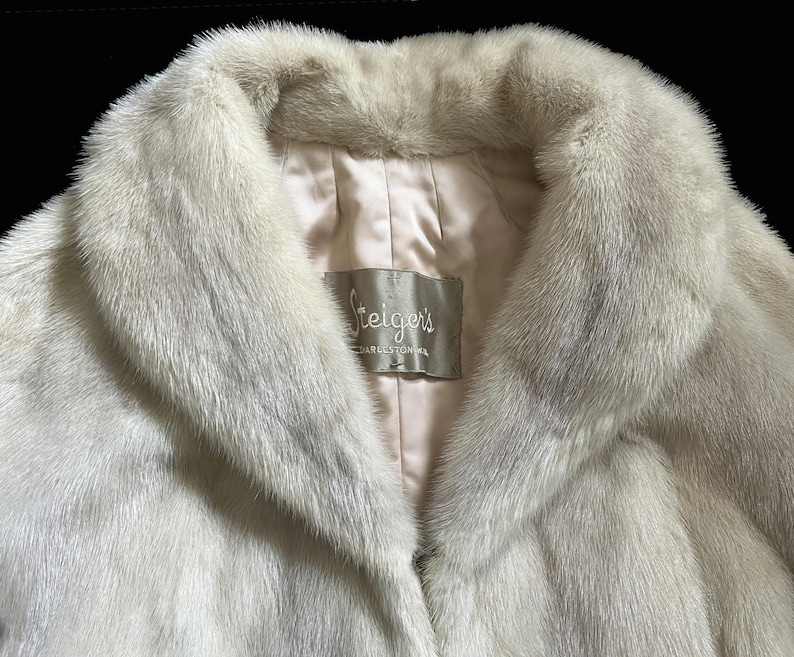 May include: A cream-coloured fur coat with a wide collar and a smooth, light-coloured satin lining. A rectangular fabric label is sewn into the interior neck area, featuring the text Steiger's and additional smaller, partially visible text.