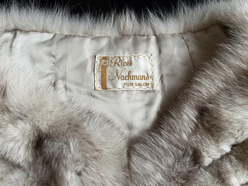 May include: Close-up of a vintage fur garment, likely a stole or collar, in shades of gray and white. The interior lining is a light cream color, with a label that reads "Rices Nachmans FUR SALON" in gold lettering.