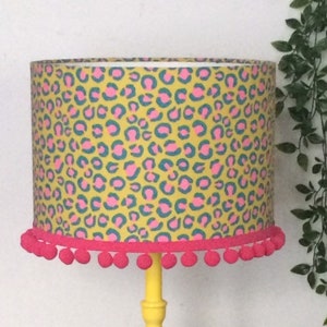 Può includere: Una lampada da tavolo gialla con un paralume a stampa leopardata rosa e blu e un bordo a pompon rosa.