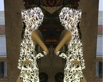 May include: Two mannequins wearing reflective black and white geometric patterned hooded dresses. The dresses have a unique, asymmetrical hemline. The background features a colorful mural and a concrete wall.