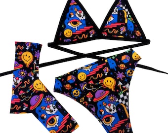 May include: A black bikini with a psychedelic print featuring smiley faces, mushrooms, and other trippy designs. The bikini top is a triangle style with adjustable straps. The bikini bottom is a high-cut style with a tie at the side.