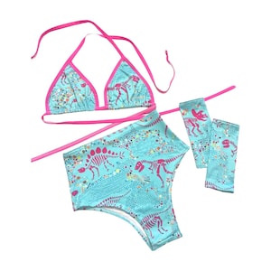 May include: A turquoise bikini set with a dinosaur skeleton print and hot pink trim. The set includes a triangle top, high-waisted bottoms, and arm sleeves. The dinosaur print is in a contrasting pink color.