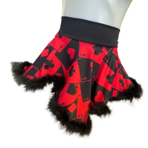 May include: A red and black skirt featuring a playing card design with hearts, clubs, diamonds, and spades. The skirt has a black waistband and is trimmed with black faux fur along the hemline. The skirt is designed to be flared.