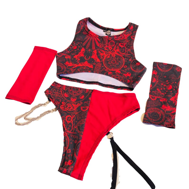 Pode incluir: Um conjunto de lingerie vermelho e preto com um top curto, roupa interior combinando e aquecedores de bra&ccedil;o. O tecido apresenta um padr&atilde;o repetido de s&iacute;mbolos e desenhos vermelhos e pretos. A roupa interior tem uma liga e uma corrente prateada.
