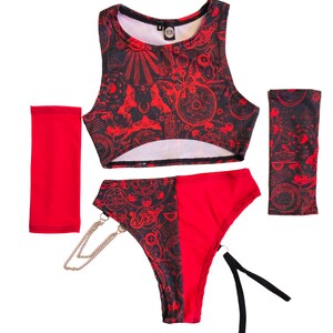 Pode incluir: Top curto vermelho e preto, roupa interior a condizer e aquecedores de bra&ccedil;o com um padr&atilde;o geom&eacute;trico vermelho e preto. O padr&atilde;o inclui c&iacute;rculos, estrelas e outras formas. A roupa interior tem uma corrente prateada presa ao lado.