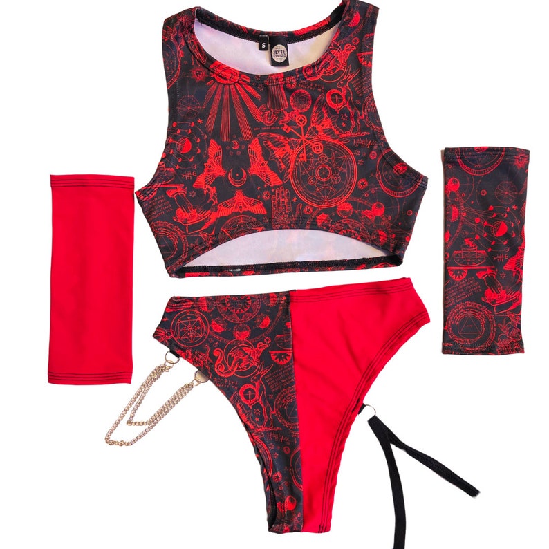 Pode incluir: Top curto vermelho e preto, roupa interior a condizer e aquecedores de bra&ccedil;o com um padr&atilde;o geom&eacute;trico vermelho e preto. O padr&atilde;o inclui c&iacute;rculos, estrelas e outras formas. A roupa interior tem uma corrente prateada presa ao lado.