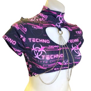 Może przedstawiać: Czarny top na ramiączkach z nadrukiem w kolorze różowym i białym z napisem "Techno" i symbolami biohazard. Top ma wycięcie w kształcie serca z przymocowanym srebrnym łańcuszkiem.