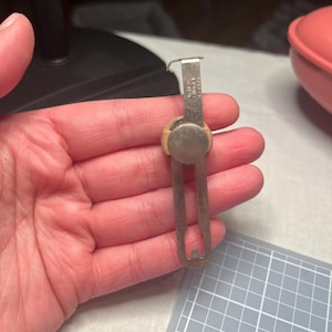 Könnte beinhalten: Ein silberfarbenes Nähmaßwerkzeug aus Metall mit einem verschiebbaren Einstellmechanismus, einem runden Knopf und einem kleinen Haken an der Oberseite. Die Metalloberfläche ist mit dem Text "MADE IN U.S.A." und "PATENT PENDING" im oberen Bereich graviert.