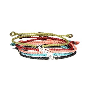 Puede incluir: Un conjunto de cinco pulseras trenzadas de colores con un dije de infinito plateado. Las pulseras están apiladas, con la pulsera verde en la parte superior y la pulsera negra en la parte inferior.