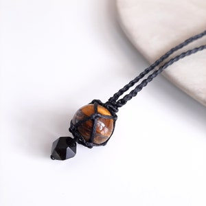 May include: A black cord necklace with a tiger's eye gemstone wrapped in a black macrame knot. A black faceted bead hangs from the bottom of the knot.