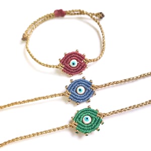 Puede incluir: Tres pulseras de cadena dorada con dijes de ojo turco de diferentes colores. Los dijes son rojo, azul y verde con ojos blancos.