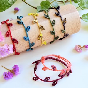 May include: A set of six colourful friendship bracelets with a leaf design. The bracelets are made with a knotted cord and are arranged on a wooden surface.