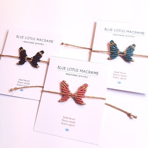 Pode incluir: Três pulseiras de macramé com desenhos de borboleta em preto, azul-turquesa e coral com detalhes dourados. Cada pulseira tem um cartão anexado com o texto "Blue Lotus Macrame" e a frase "kind heart, fierce mind, brave spirit".