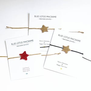 Può includere: Tre braccialetti a forma di stella in macramè con stelle di diversi colori. I braccialetti sono su delle carte con il testo "Blue Lotus Macrame - macrame jewelry -" e "Buon Natale e Felice Anno Nuovo".