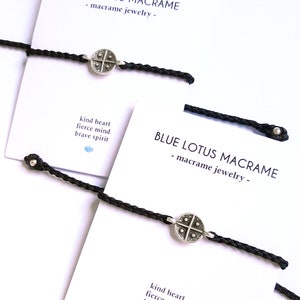 Può includere: Due braccialetti in macramè neri con ciondoli argentati. I ciondoli hanno un design a croce e sono attaccati ai braccialetti con un anello d'argento. I braccialetti sono esposti su carte bianche con il testo "BLUE LOTUS MACRAME - macramè gioielli -" e "kind heart fierce mind brave spirit".