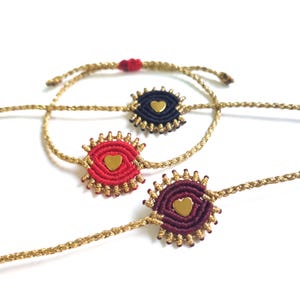 May include: Three friendship bracelets with gold cord and a small gold heart charm in the center of each circle. The circles are made of red, black, and burgundy thread.