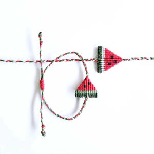 Puede incluir: Pulsera de macramé con dos dijes de rebanadas de sandía. Los dijes son rojos con semillas negras y corteza verde y blanca. El cordón de la pulsera es rojo, verde y blanco, con un cierre de nudo corredizo.