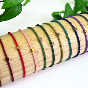 Könnte beinhalten: Ein Set aus 12 geflochtenen Armbändern mit herzförmigen Anhängern in verschiedenen Farben, darunter Rot, Rosa, Grün, Blau und Lila. Die Armbänder werden auf einer Holzoberfläche präsentiert.