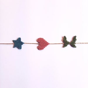 May include: A string of macrame knots with a blue star, pink heart, and green bow. The knots are strung on a natural twine.