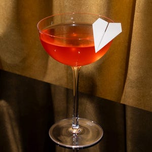 May include: A clear glass coupe cocktail glass filled with a vibrant orange-red liquid. A small, white, folded paper airplane garnish is perched on the rim of the glass. The stem is slender and leads to a circular base, holding the drink upright.