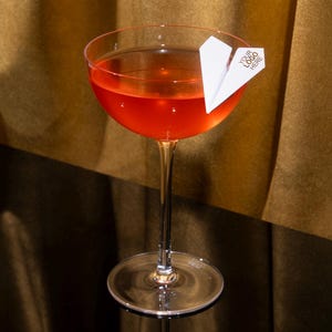 Könnte beinhalten: Ein klares Cocktailschale aus Glas, gefüllt mit einer leuchtend orange-roten Flüssigkeit. Eine kleine Garnitur aus weißem Papier in Form eines Papierfliegers sitzt am Rand des Glases und trägt den Aufdruck YOUR LOGO HERE in schwarzen Großbuchstaben.