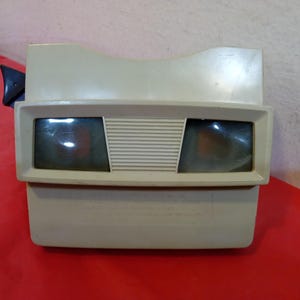 Könnte beinhalten: Ein Vintage-View-Master-Betrachter in Beige. Der Betrachter hat zwei Okulare, einen zentralen rechteckigen Abschnitt mit horizontalen Linien und einen schwarzen Hebel an der Seite. Das Gerät befindet sich auf einer roten Oberfläche.