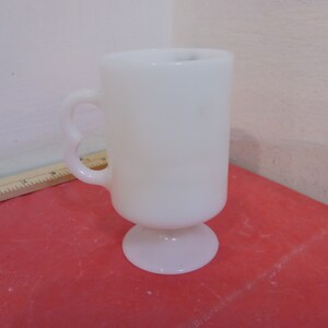 May include: A white ceramic mug with a handle and a pedestal base. The mug is on a red surface.