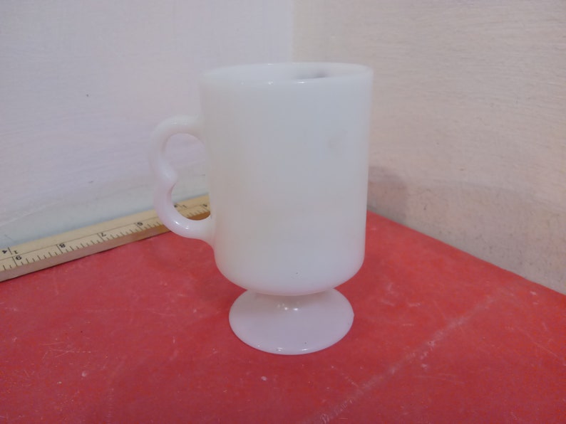 May include: A white ceramic mug with a handle and a pedestal base. The mug is on a red surface.