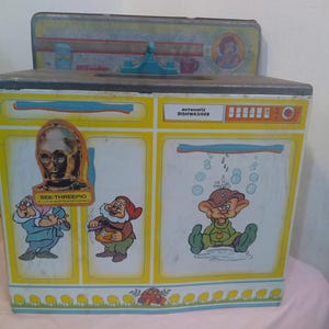May include: A vintage toy dishwasher with a yellow and white design. The dishwasher has a blue and white stripe across the top and a red button on the right side. The front of the dishwasher features three cartoon characters: a dwarf with a red hat, a dwarf with a green hat, and a robot. The text "Automatic Dishwasher" is printed on the front of the dishwasher.