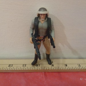 May include: A Star Wars action figure of a Rebel soldier wearing a white helmet and a brown vest. The figure is holding a black blaster rifle in one hand and a black blaster pistol in the other hand. The figure is standing on a wooden ruler.