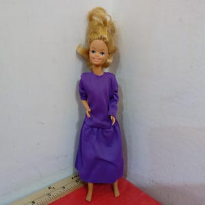May include: A vintage doll with blonde hair and blue eyes, wearing a long-sleeved purple dress. The doll is standing upright, showcasing the full length of the outfit. The doll's hair is styled in a voluminous, layered fashion.