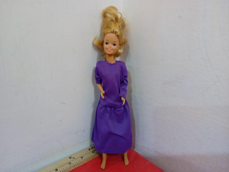 May include: A vintage doll with blonde hair and blue eyes, wearing a long-sleeved purple dress. The doll is standing upright, showcasing the full length of the outfit. The doll's hair is styled in a voluminous, layered fashion.