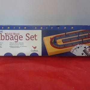 Op de afbeelding: Deluxe Cribbage Set in een blauwe doos. De doos heeft de tekst "Premier Edition" en "Deluxe Cribbage Set". De set bevat een massief houten bord, pinnen en Bicycle speelkaarten. Het bord heeft een doorlopende baan.
