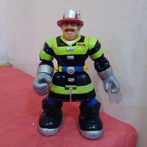 May include: A firefighter action figure with a red helmet, black and green uniform, and a silver tool belt. The figure has a mustache and is holding a silver tool in one hand.