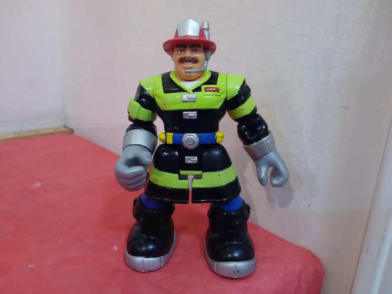 May include: A firefighter action figure with a red helmet, black and green uniform, and a silver tool belt. The figure has a mustache and is holding a silver tool in one hand.