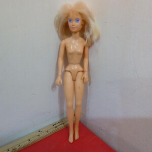 May include: A nude-colored fashion doll with blonde hair and blue eyes. The doll is standing on a red surface, with a ruler partially visible in the foreground. The doll's arms and legs are articulated.