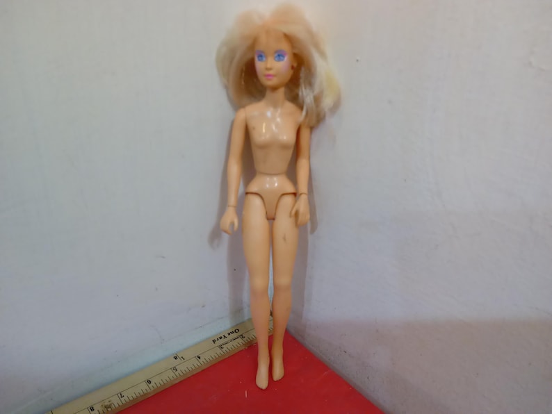 May include: A nude-colored fashion doll with blonde hair and blue eyes. The doll is standing on a red surface, with a ruler partially visible in the foreground. The doll's arms and legs are articulated.