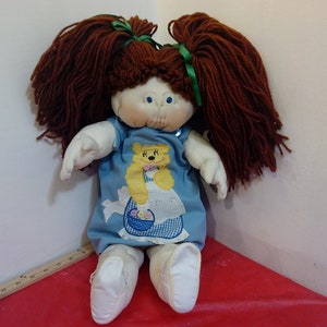 May include: A vintage Cabbage Patch Kid doll with brown yarn hair in pigtails, wearing a blue dress with a yellow embroidered bear design.
